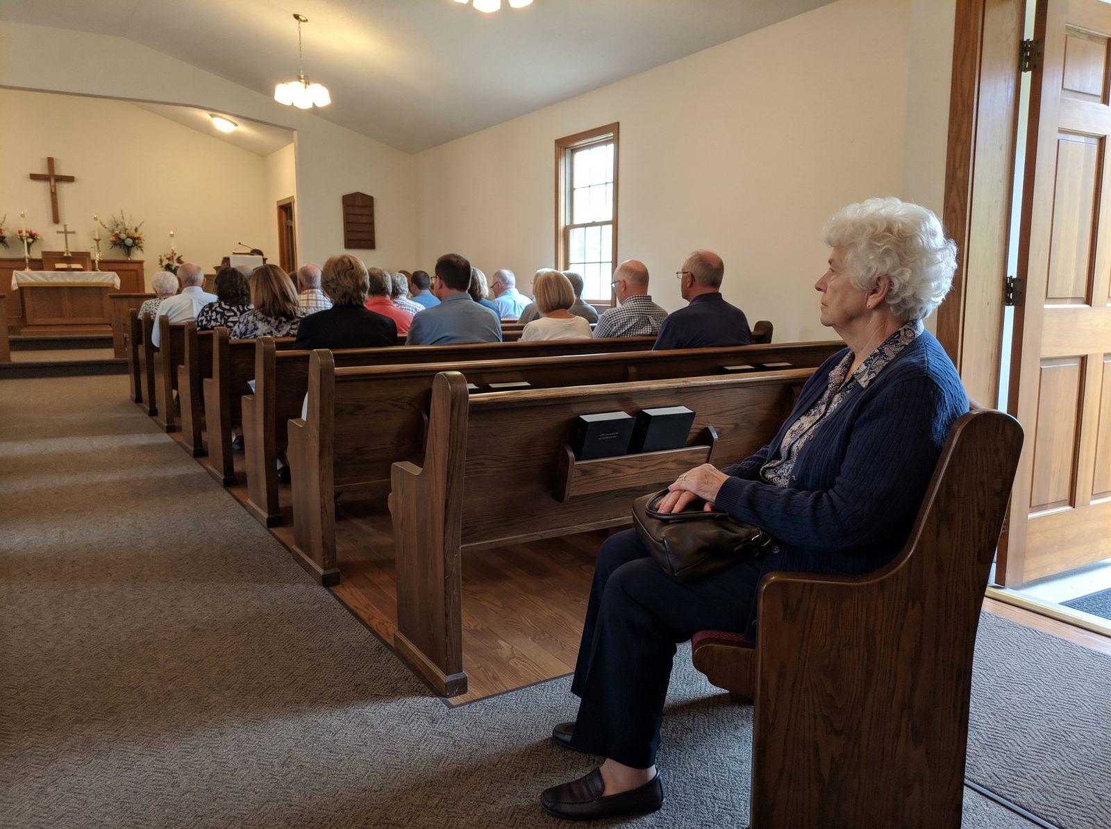 An older woman with white hair sits alone at the end of a back pew near the church door