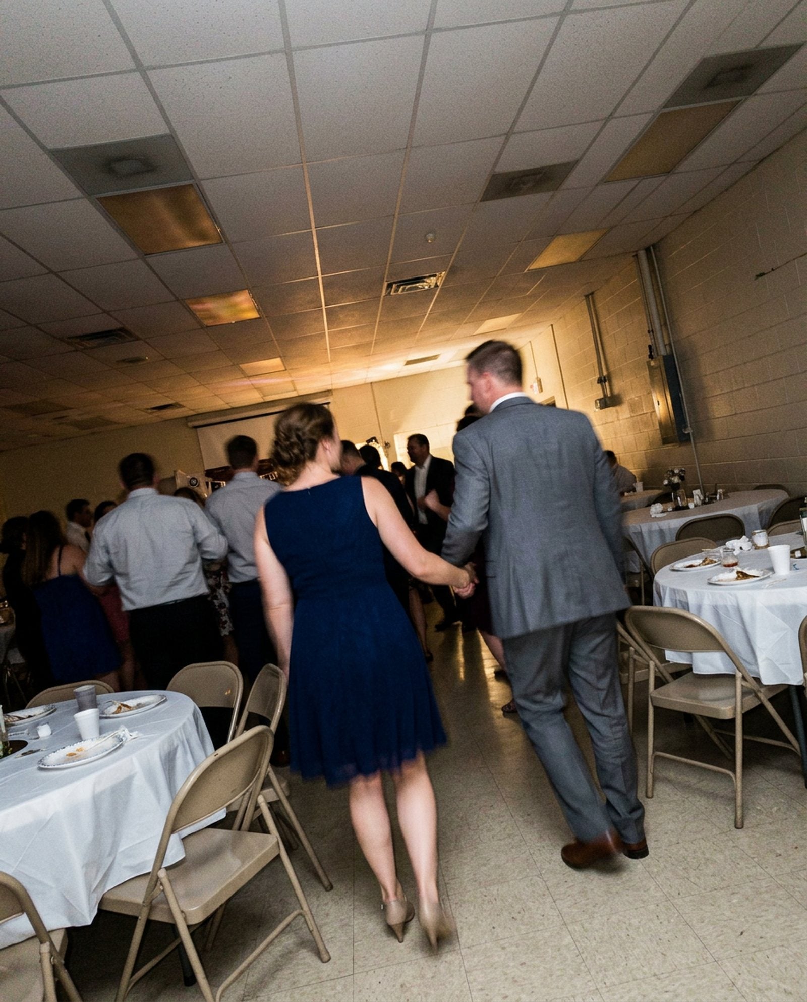 A woman in a navy dress and a man in a grey suit walking hand in hand toward the dance floor at a wedding reception, seen from behind, between round tables and folding chairs